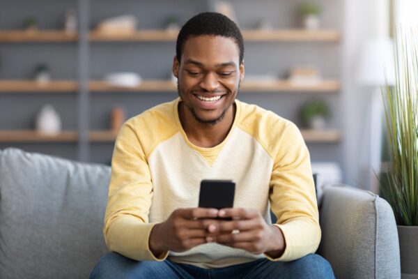 Smiling,Black,Guy,Sitting,On,Couch,At,Home,,Using,Smartphone,